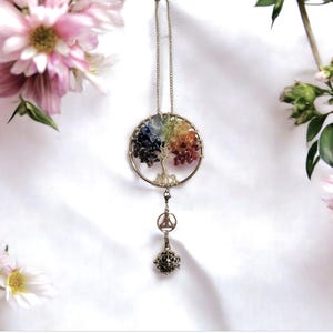 May include: A silver-toned tree of life pendant with a rainbow of gemstones representing the leaves. The pendant hangs from a chain and features a charm with a small bell. The background is white with pink flowers.
