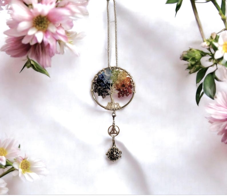 May include: A silver-toned tree of life pendant with a rainbow of gemstones representing the leaves. The pendant hangs from a chain and features a charm with a small bell. The background is white with pink flowers.