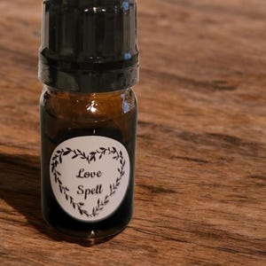 May include: A small, brown glass bottle with a black cap. The bottle has a white label with the words "Love Spell" inside a heart-shaped wreath. The bottle is filled with a dark liquid.