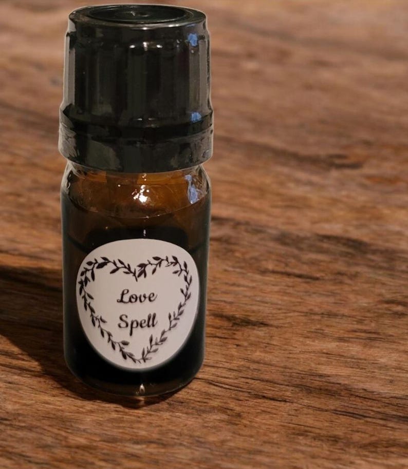 May include: A small, brown glass bottle with a black cap. The bottle has a white label with the words "Love Spell" inside a heart-shaped wreath. The bottle is filled with a dark liquid.