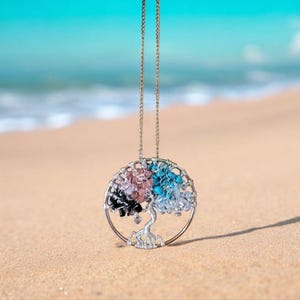Puede incluir: Un collar plateado con un colgante de &aacute;rbol de la vida. El colgante presenta un dise&ntilde;o de &aacute;rbol con alambre con cuentas de colores en negro, rosa, azul y transparente. El collar se muestra en una playa de arena con el mar de fondo.