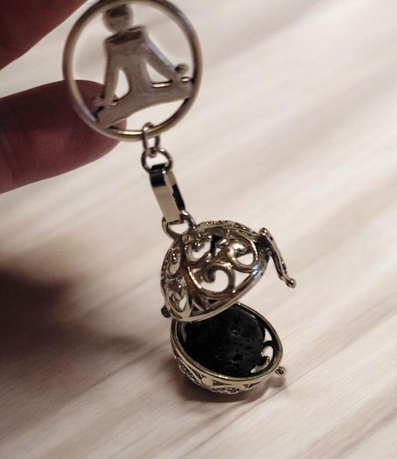 May include: Silver aromatherapy pendant with a filigree design, featuring a round charm with a seated figure. The pendant opens to reveal a black lava stone. A stylish accessory for essential oils.