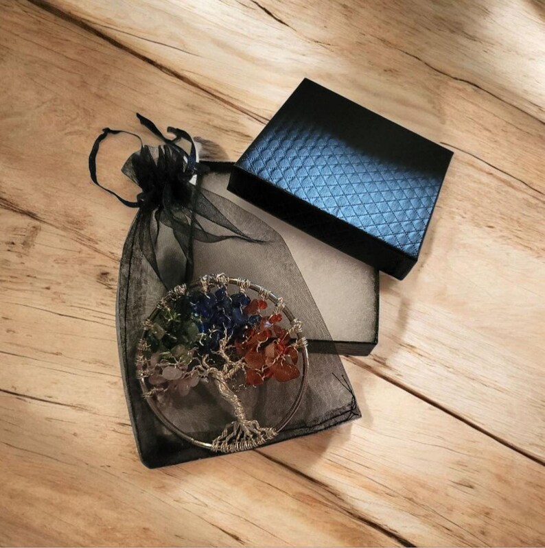 Puede incluir: Una escultura de &aacute;rbol de la vida de alambre con piedras preciosas coloridas, exhibida en una bolsa de organza negra, junto a una caja de regalo negra texturizada con interior azul. El &aacute;rbol presenta un marco de alambre plateado y piedras de colores.