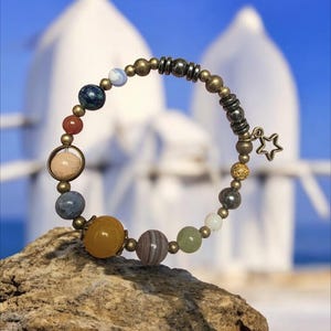 May include: A beaded bracelet featuring colorful gemstone beads representing planets, connected by gold-toned metal beads. A small star charm hangs from the bracelet. The bracelet is displayed on a rock with a blurred background of white buildings and a blue sky.