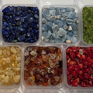 May include: Assortment of gemstones in clear plastic containers. The gemstones are in various colors, including purple, blue, light blue, green, yellow, brown, and red. The containers are arranged in a grid pattern.