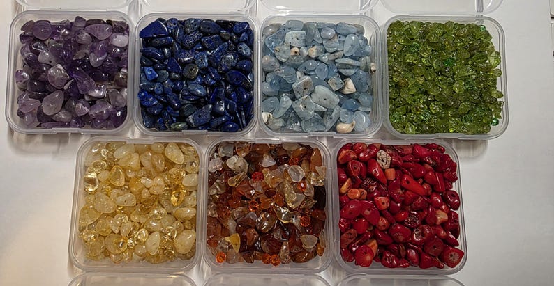 May include: Assortment of gemstones in clear plastic containers. The gemstones are in various colors, including purple, blue, light blue, green, yellow, brown, and red. The containers are arranged in a grid pattern.