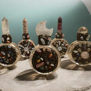 May include: Five clear glass perfume bottles with decorative tops and filled with colorful stones. Each bottle has a different crystal or stone top, including a clear quartz point, a pink stone, a moon-shaped crystal, and a striped crystal.