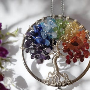 May include: A silver-toned pendant necklace featuring a tree of life design. The tree's branches and leaves are crafted from colorful gemstones, including red, orange, yellow, green, blue, and purple. The pendant hangs from a delicate silver chain.