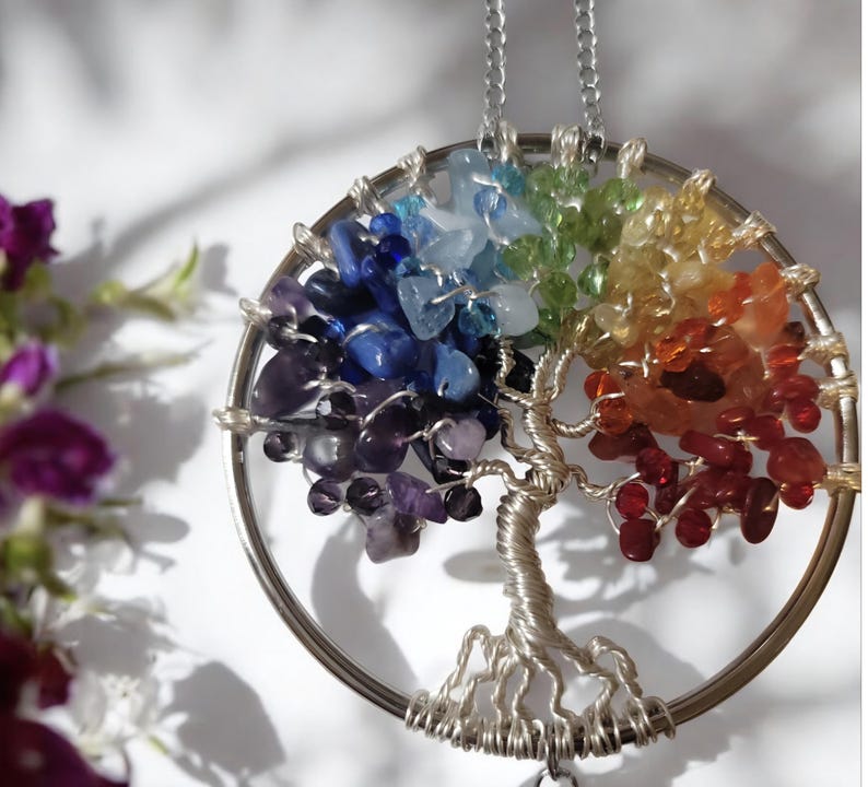 May include: A silver-toned pendant necklace featuring a tree of life design. The tree's branches and leaves are crafted from colorful gemstones, including red, orange, yellow, green, blue, and purple. The pendant hangs from a delicate silver chain.