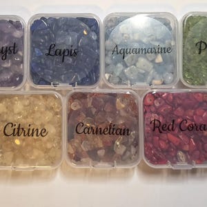 May include: Seven small, clear plastic containers filled with various gemstones. The containers are labeled with the names of the stones: Amethyst, Lapis, Aquamarine, Peridot, Citrine, Carnelian, and Red Coral. The gemstones are in shades of purple, blue, green, yellow, orange, and red.