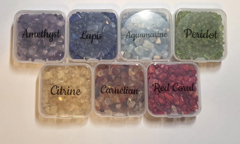 May include: Seven small, clear plastic containers filled with various gemstones. The containers are labeled with the names of the stones: Amethyst, Lapis, Aquamarine, Peridot, Citrine, Carnelian, and Red Coral. The gemstones are in shades of purple, blue, green, yellow, orange, and red.