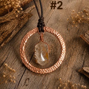 May include: A necklace featuring a hammered copper circular pendant suspended from a dark brown leather cord. A clear, hexagonal crystal with golden inclusions is wrapped in copper wire and hangs from the center of the copper ring. Text #2.