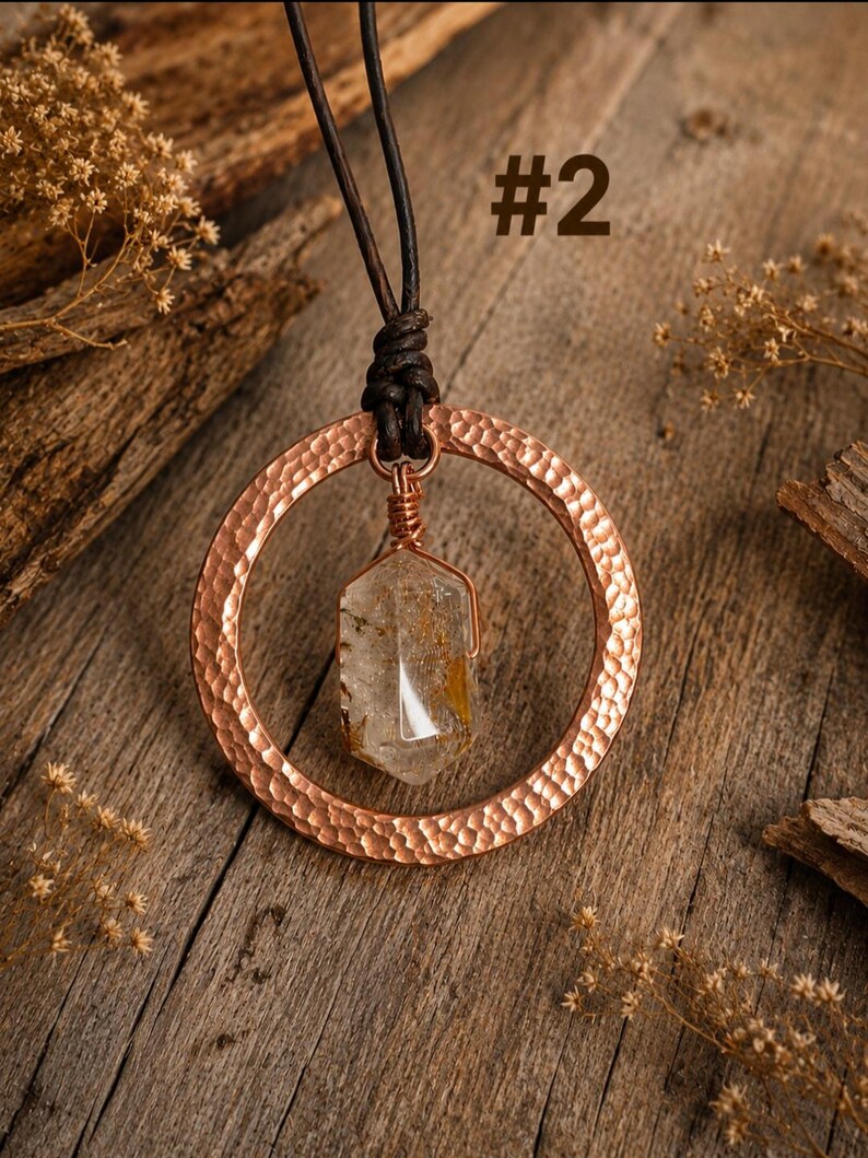 May include: A necklace featuring a hammered copper circular pendant suspended from a dark brown leather cord. A clear, hexagonal crystal with golden inclusions is wrapped in copper wire and hangs from the center of the copper ring. Text #2.