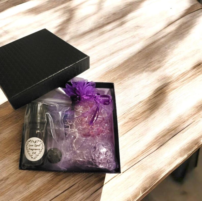 May include: A black gift box with the lid slightly open, revealing a bottle of "Love Spell Fragrance Oil", a small black stone, and a purple sachet. The box is on a wooden surface.