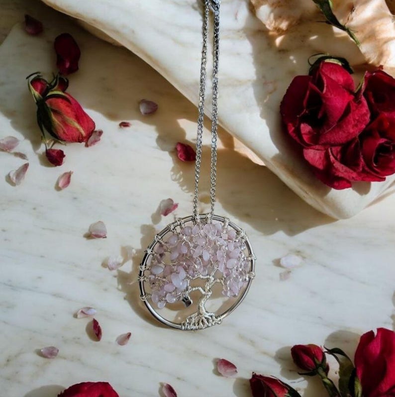 Puede incluir: Un collar plateado con un colgante circular que representa un &aacute;rbol de la vida. Las hojas del colgante est&aacute;n hechas de peque&ntilde;as piedras de color rosa p&aacute;lido. El collar se exhibe sobre una superficie de m&aacute;rmol, rodeado de rosas rojas y p&eacute;talos rosas.