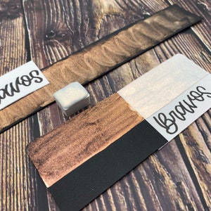 May include: A close-up of a small, white, square container of metallic gold paint with a black and gold paint swatch next to it. The paint swatch has the word "Bravos" written on it.