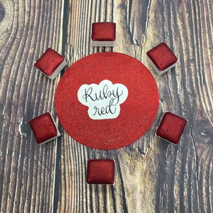 May include: A collection of ruby red watercolor paint pans arranged around a red glitter circle with the words "Ruby red" in white script. The paint pans are square and set in white containers, on a wooden background.