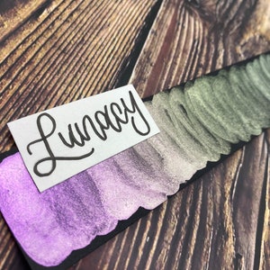 May include: A close-up of a black card with swatches of metallic paint in shades of purple, silver, and green. The card is labeled "Lunacy" in cursive.