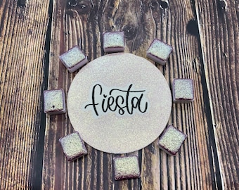 May include: A white circle with the word "fiesta" written in black script. The circle is surrounded by eight small, square, iridescent tiles with a gold and purple shimmer.