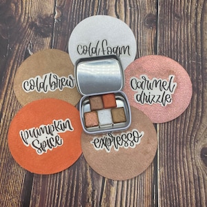 May include: A set of five round, metallic coasters with different fall-themed designs. The coasters feature the words "cold brew", "cold foam", "caramel drizzle", "pumpkin spice", and "espresso".