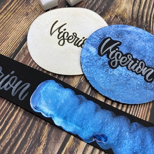 May include: Two white coasters with the word "Viserion" written in black ink. A black leather strip with a blue metallic paint swipe.