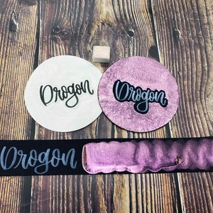 May include: Two white circles with the word "Drogon" written in black ink. A black rectangle with the word "Drogon" written in silver ink and a purple metallic paint swatch.