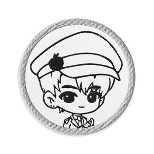 Op de afbeelding: Geborduurde patch met een stripfiguur met een hoed en uniform in militaire stijl. Het ontwerp is in zwarte draad op een witte cirkelvormige achtergrond met een grijs gestikte rand. Het karakter heeft grote ogen en een kleine glimlach.