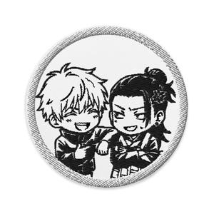 Op de afbeelding: Een ronde geborduurde patch met een zwart-witte grafische illustratie van twee anime-stijl personages. Het ene personage heeft stekelig haar en een brede glimlach, terwijl de andere donker haar in een staart en een grijns heeft. De patch heeft een rand.