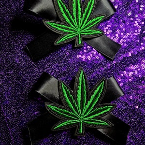 以下が含まれることがあります： 黒いサテンリボンの蝶結びが2つ。それぞれに緑色の大麻の葉のパッチが付いています。パッチは詳細な葉のデザインで刺繍されています。背景はキラキラ光る紫色のスパンコール生地です。