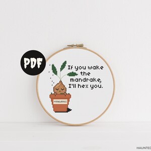 Op de afbeelding: Een witte borduurring met een kruissteekontwerp van een mandragora-plant in een pot. De tekst "Als je de mandragora wakker maakt, betover ik je." is boven de plant geborduurd.