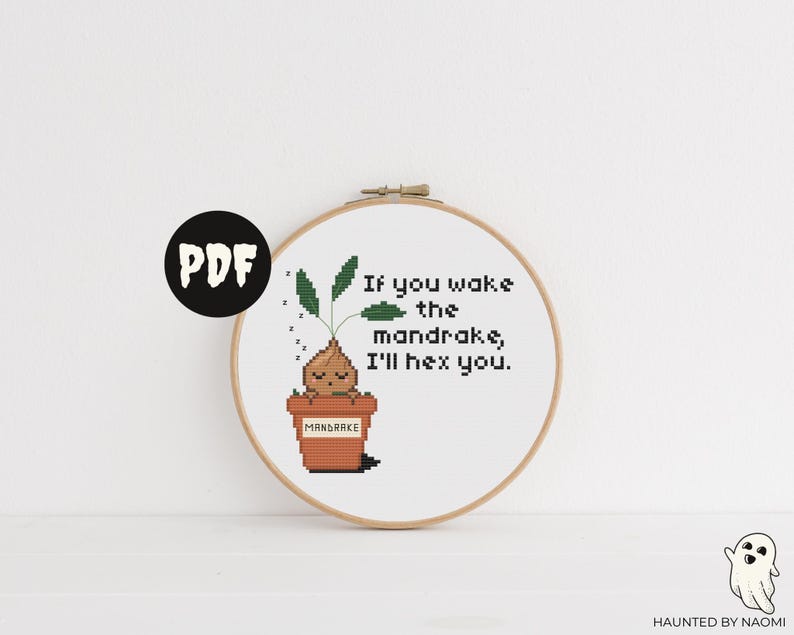 Op de afbeelding: Een witte borduurring met een kruissteekontwerp van een mandragora-plant in een pot. De tekst "Als je de mandragora wakker maakt, betover ik je." is boven de plant geborduurd.