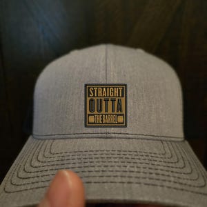Op de afbeelding: Een grijze en zwarte trucker-pet met een zwart en gouden patch waarop "STRAIGHT OUTTA THE BARREL" staat.
