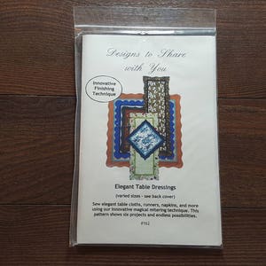 May include: A white paper pattern booklet with a blue and green design on the front cover. The title is "Designs to Share with You" and the subtitle is "Elegant Table Dressings". The booklet is for sewing tablecloths, runners, and napkins.