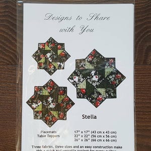 Può includere: Un pacchetto di modelli di trapuntatura chiamato "Stella" con disegni floreali a forma di stella. Include tovagliette e copritavolo in tre misure: 43 cm x 43 cm, 56 cm x 56 cm e 66 cm x 66 cm. Il testo "Designs to Share with You" è in alto.