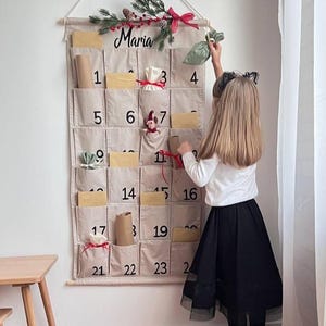 Peut inclure: Un calendrier de l'Avent mural en tissu beige avec vingt-quatre poches numérotées, portant le prénom Maria en écriture noire en haut. Le calendrier est décoré d'une branche de pin, de baies rouges et d'un ruban rouge, contenant des petits cadeaux.