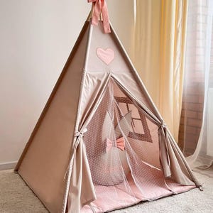 May include: A blush pink teepee tent with a heart applique and decorative accents. The tent features a mesh window, a pink polka dot interior, and a matching pink floor mat. The top is adorned with fabric leaves and a bow.