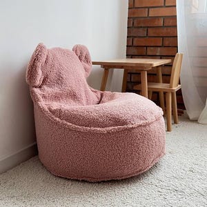 May include: A pink teddy bear-shaped beanbag chair with ear-like protrusions on the backrest. The chair is made of a soft, textured material and sits on a light-coloured carpet. A small wooden table and chair are in the background.