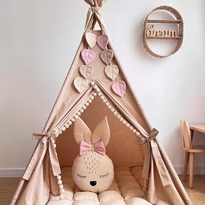 Op de afbeelding: Een beige tipi-tent met een konijnenkussen en bladvormige decoraties. De tent heeft een zachte, gewatteerde vloer en is versierd met pompons. Een houten plank met de naam "Shalom" staat aan de muur.