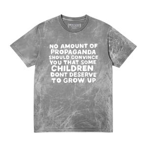May include: A grey acid-wash t-shirt with a crew neckline and short sleeves. The front features white text that reads: NO AMOUNT OF PROPAGANDA SHOULD CONVINCE YOU THAT SOME CHILDREN DONT DESERVE TO GROW UP. The fabric has a mottled, marbled texture.