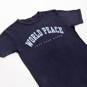 K&ouml;nnte beinhalten: Marineblaues T-Shirt mit dem Schriftzug "WORLD PEACE" in wei&szlig;er, bogenf&ouml;rmiger Schrift mit hellblauer Umrandung. Darunter sind die Worte "LOVE EACH OTHER" in Wei&szlig; gedruckt. Das T-Shirt ist aus weichem Material.