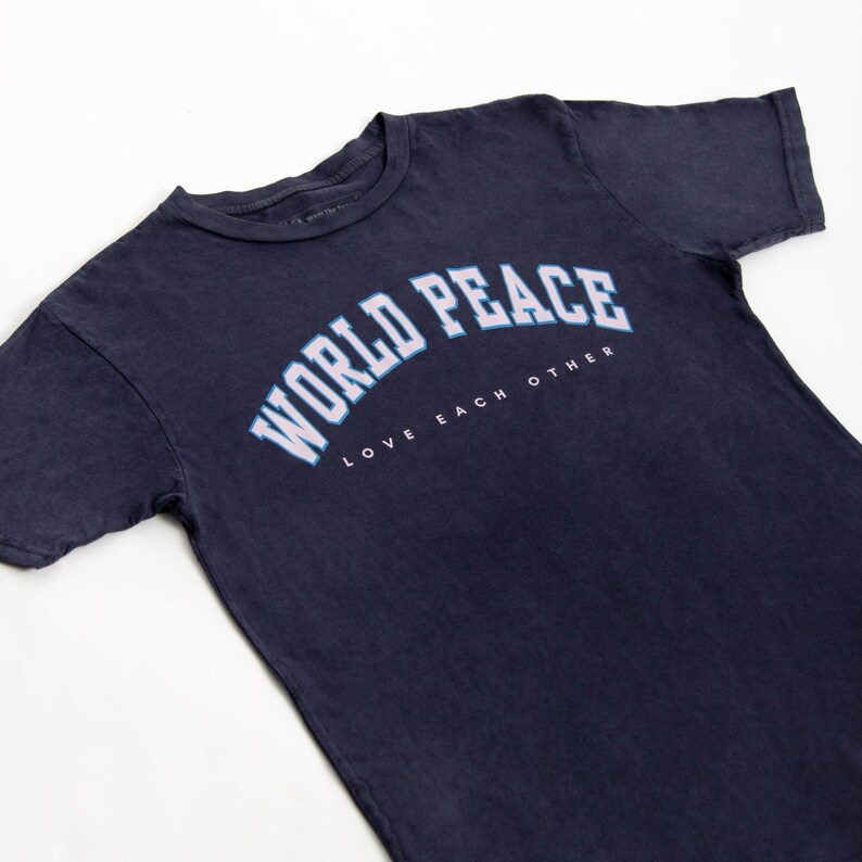 K&ouml;nnte beinhalten: Marineblaues T-Shirt mit dem Schriftzug "WORLD PEACE" in wei&szlig;er, bogenf&ouml;rmiger Schrift mit hellblauer Umrandung. Darunter sind die Worte "LOVE EACH OTHER" in Wei&szlig; gedruckt. Das T-Shirt ist aus weichem Material.