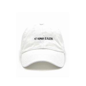 May include: A white baseball cap featuring the embroidered black text NO HUMAN IS ILLEGAL centred on the front panel. The cap has a structured crown, a curved brim, and a button at the top, constructed from a solid white fabric material.