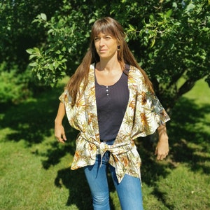 Könnte beinhalten: Eine Frau trägt einen weißen und braunen floralen Kimono mit einem schwarzen Oberteil und einer blauen Jeans. Der Kimono hat einen Gürtel in der Taille und ist vorne offen.