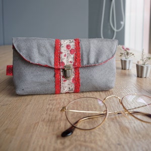 Può includere: Un astuccio per occhiali in tessuto grigio con un bordo floreale rosso e bianco e una chiusura in metallo. L'astuccio è appoggiato su una superficie in legno accanto a un paio di occhiali con montatura dorata.