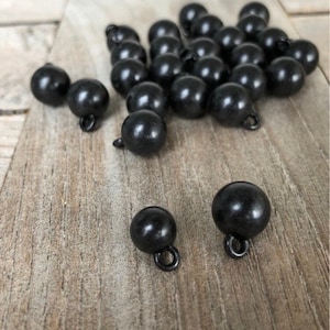 Può includere: Un gruppo di bottoni rotondi neri con un piccolo anello sul fondo. I bottoni sono sparsi su una superficie di legno.