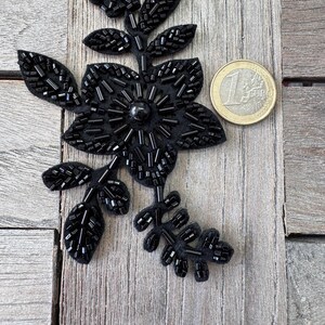 May include: A black beaded floral applique with a flower and leaves design. The applique is embellished with small, black beads. A Euro coin is next to the applique for size reference.