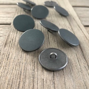Puede incluir: Un conjunto de diez botones redondos de metal gris oscuro con un bucle en la parte posterior. Los botones están esparcidos sobre una superficie de madera.
