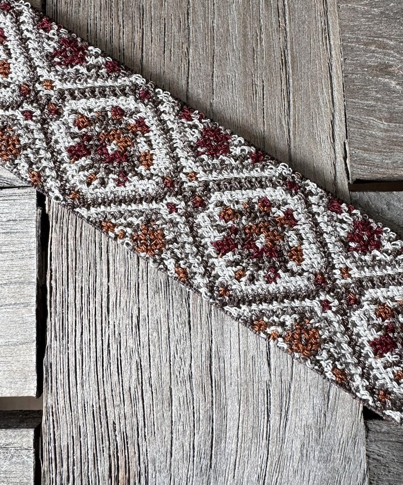May include: Close-up of a crocheted textile with a geometric pattern. The design features diamond shapes in white, brown, and burgundy. The textile is placed on a weathered wooden surface, showcasing the intricate details of the crochet work.