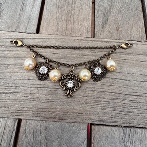 K&ouml;nnte beinhalten: Ein antikes goldfarbenes Armband mit einer Kette und dekorativen Anh&auml;ngern. Die Anh&auml;nger umfassen Kunstperlen, klare Strasssteine und verzierte Filigranmuster. Das Armband hat zwei Verschlussverschl&uuml;sse.