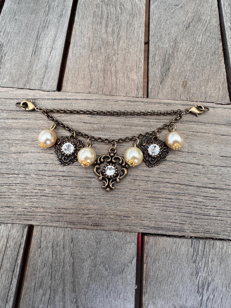 K&ouml;nnte beinhalten: Ein antikes goldfarbenes Armband mit einer Kette und dekorativen Anh&auml;ngern. Die Anh&auml;nger umfassen Kunstperlen, klare Strasssteine und verzierte Filigranmuster. Das Armband hat zwei Verschlussverschl&uuml;sse.